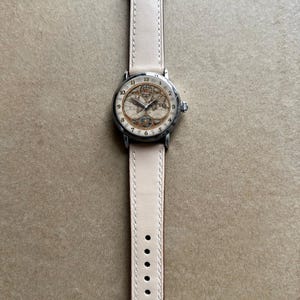 May include: A wristwatch with a cream-colored leather band and a silver-toned case. The watch face features a vintage map design with black numerals. The band is approximately 0.75 inches wide.