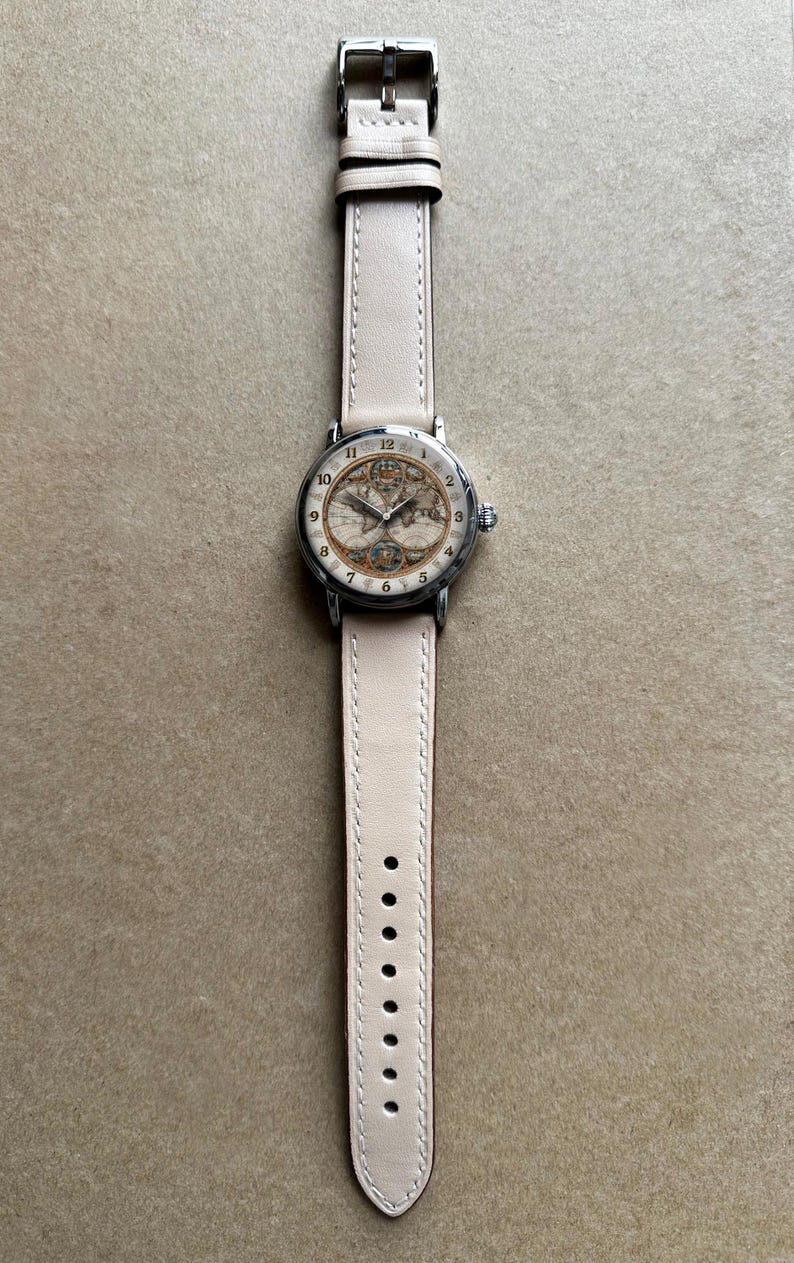 May include: A wristwatch with a cream-colored leather band and a silver-toned case. The watch face features a vintage map design with black numerals. The band is approximately 0.75 inches wide.