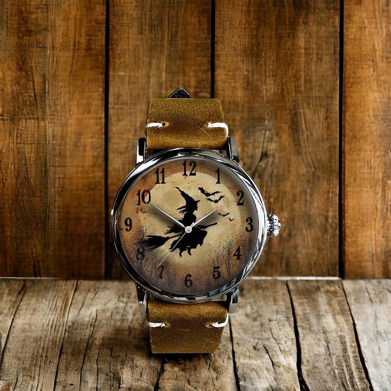 May include: A wristwatch with a brown leather band and a silver-colored case. The watch face features a Halloween-themed design with a witch on a broomstick, a full moon, and bats. The watch has a classic analog display.