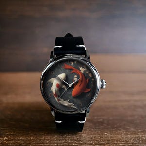 Op de afbeelding: Ronde polshorloge met een zwarte leren band. De wijzerplaat toont twee koi-karpers, &eacute;&eacute;n rood en &eacute;&eacute;n wit, tegen een donkergrijze achtergrond. Het horloge heeft zilverkleurige wijzers en een zilverkleurige lunette.