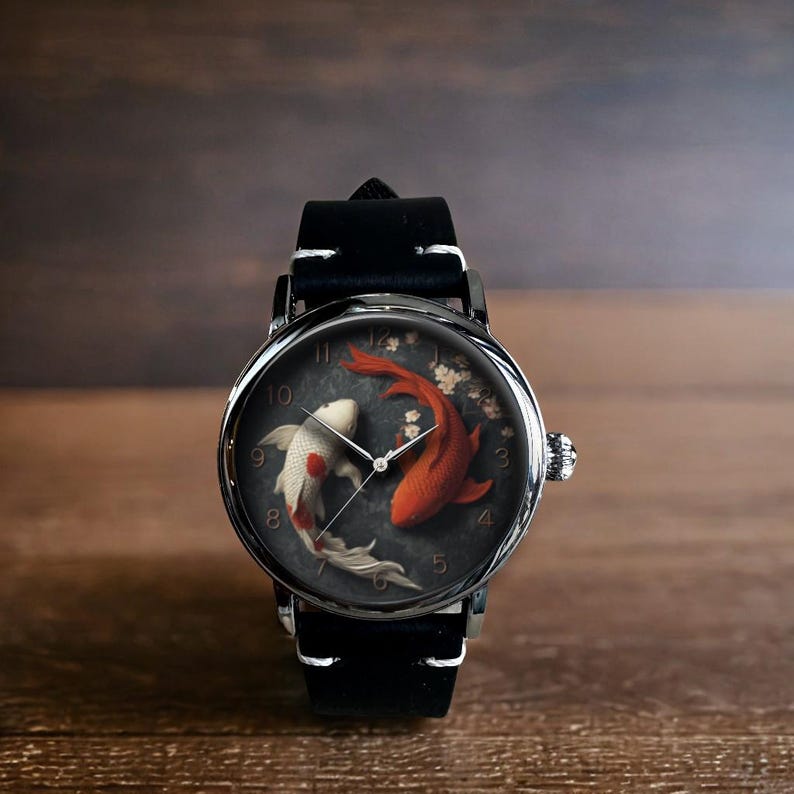 Op de afbeelding: Ronde polshorloge met een zwarte leren band. De wijzerplaat toont twee koi-karpers, &eacute;&eacute;n rood en &eacute;&eacute;n wit, tegen een donkergrijze achtergrond. Het horloge heeft zilverkleurige wijzers en een zilverkleurige lunette.