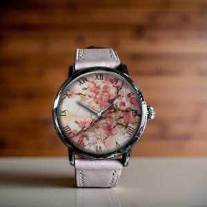 Peut inclure: Une montre-bracelet avec un boîtier argenté et un bracelet en cuir rose clair. Le cadran de la montre présente un motif floral avec des fleurs roses et des chiffres romains. La montre a un style classique et élégant.