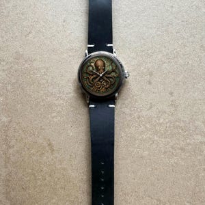 May include: A round wristwatch with a black leather band. The watch face features a detailed illustration of an octopus in shades of brown and green. The watch has a silver-colored case and a black leather strap.