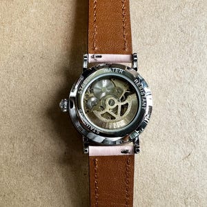May include: A silver wristwatch with a brown leather band. The watch face is clear and shows the inner workings of the watch. The watch face is labeled 'STAINLESS STEEL' and 'WATER RESISTANT'.