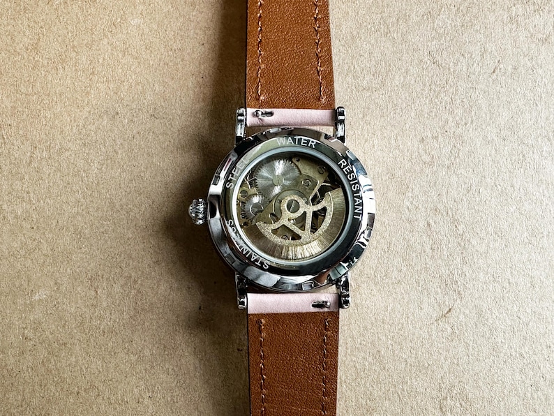May include: A silver wristwatch with a brown leather band. The watch face is clear and shows the inner workings of the watch. The watch face is labeled 'STAINLESS STEEL' and 'WATER RESISTANT'.