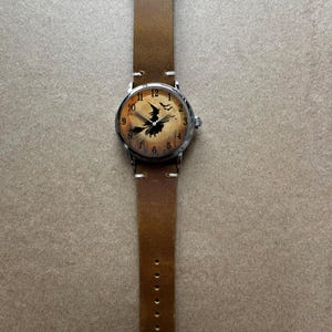 May include: A brown leather watch band with a silver watch face. The watch face features a witch on a broomstick design. The watch has a brown leather band with a buckle closure. The watch face has the numbers 1 through 12.