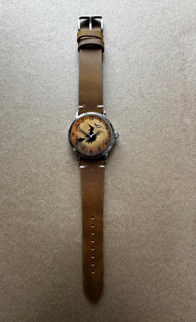 May include: A brown leather watch band with a silver watch face. The watch face features a witch on a broomstick design. The watch has a brown leather band with a buckle closure. The watch face has the numbers 1 through 12.