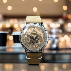May include: A wristwatch with a silver-toned circular case and a tan leather strap. The watch face features a detailed sepia-toned illustration of a sailing ship on the ocean, surrounded by an ornate decorative border and thin silver hands.