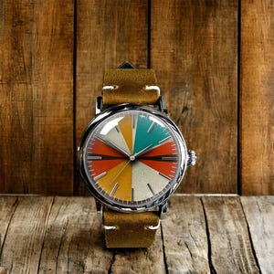 Op de afbeelding: Een ronde polshorloge met een kleurrijke, gesegmenteerde wijzerplaat in oranje, turquoise, geel en cr&egrave;me. Het horloge heeft een zilveren kast en een bruine leren band. De wijzerplaat heeft een uniek ontwerp met een retro-esthetiek.