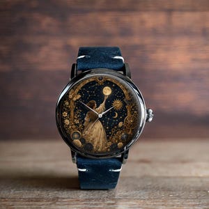 May include: A round wristwatch with a black case and a navy blue leather band. The watch face features a celestial design with a woman reaching for the moon, surrounded by stars and golden floral accents. The watch has a silver crown.