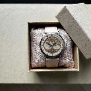 May include: A wristwatch with a cream-colored leather band and a round face featuring a vintage map design. The watch is presented in a small, open cardboard box with a burlap-textured cushion.