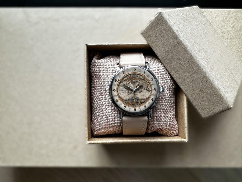 May include: A wristwatch with a cream-colored leather band and a round face featuring a vintage map design. The watch is presented in a small, open cardboard box with a burlap-textured cushion.
