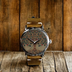 Puede incluir: Un reloj de pulsera con caja plateada y correa de cuero marrón claro. La esfera del reloj presenta una ilustración detallada de un árbol con hojas verdes y detalles rojos. El reloj está sobre un fondo de madera.