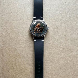 May include: A round wristwatch with a black leather band. The watch face features an octopus illustration in shades of brown and green. The watch has a silver-colored case and a black leather strap with white stitching.