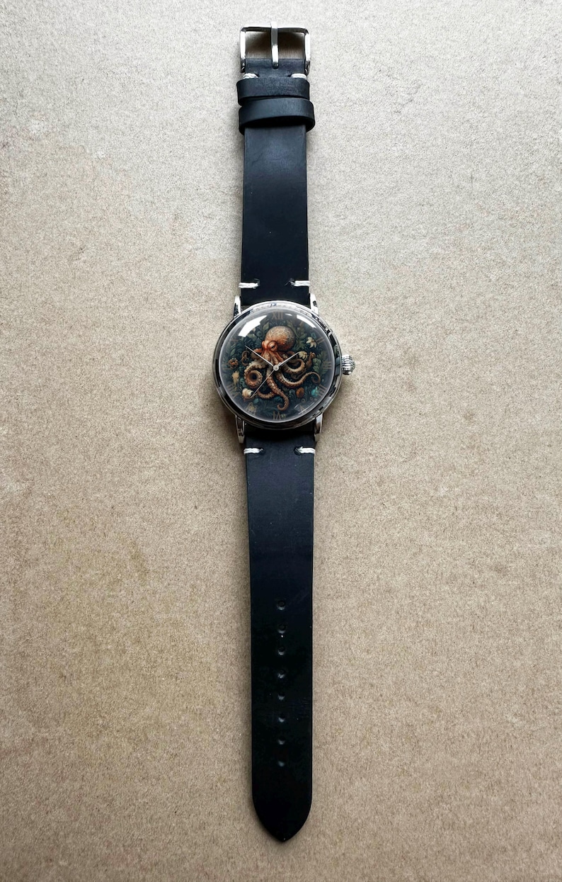 May include: A round wristwatch with a black leather band. The watch face features an octopus illustration in shades of brown and green. The watch has a silver-colored case and a black leather strap with white stitching.