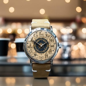 Puede incluir: Un reloj de pulsera plateado con una correa de cuero beige. La esfera del reloj presenta un dise&ntilde;o detallado del zod&iacute;aco con s&iacute;mbolos astrol&oacute;gicos y un centro azul oscuro. El reloj tiene un estilo cl&aacute;sico y elegante.