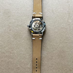 Puede incluir: Un reloj de pulsera con una correa de cuero marr&oacute;n claro y una caja de acero inoxidable. La esfera del reloj es parcialmente transparente, revelando el mecanismo interno. La correa tiene costuras blancas y una hebilla plateada. El reloj es resistente al agua.