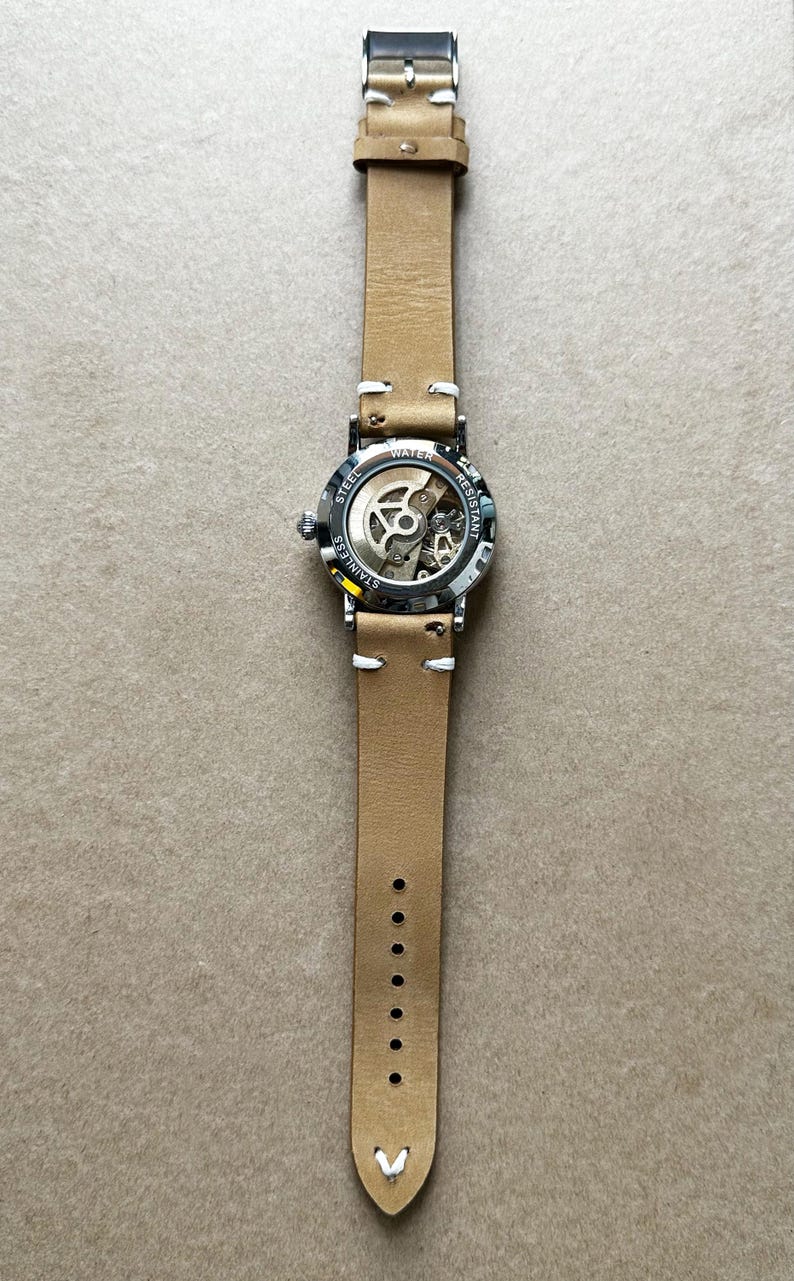 Puede incluir: Un reloj de pulsera con una correa de cuero marr&oacute;n claro y una caja de acero inoxidable. La esfera del reloj es parcialmente transparente, revelando el mecanismo interno. La correa tiene costuras blancas y una hebilla plateada. El reloj es resistente al agua.