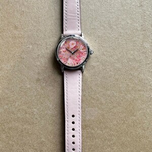 May include: A wristwatch with a pale pink leather strap and a silver-coloured case. The watch face displays a floral design with pink blossoms. The watch is shown on a neutral-toned surface.