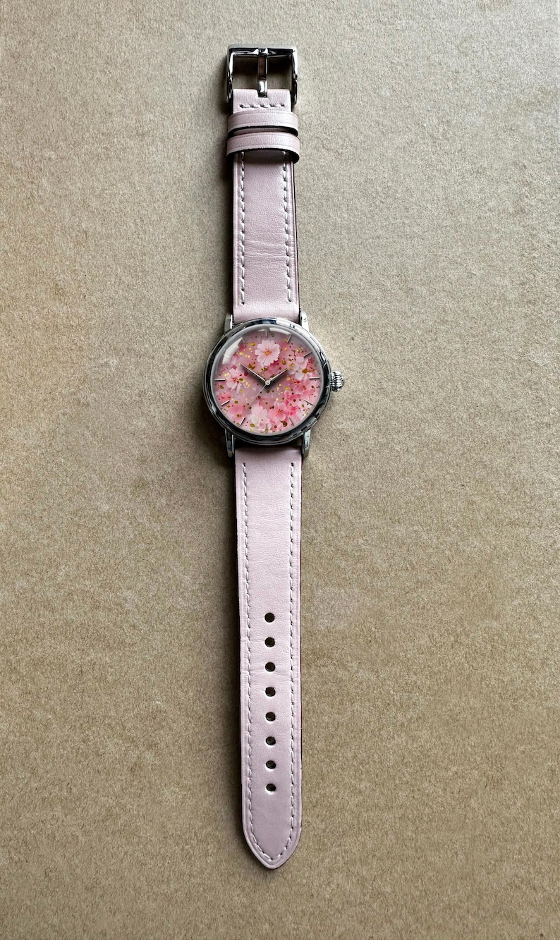 May include: A wristwatch with a pale pink leather strap and a silver-coloured case. The watch face displays a floral design with pink blossoms. The watch is shown on a neutral-toned surface.
