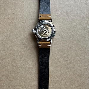 May include: A stainless steel wristwatch with a skeleton dial, showcasing the inner workings. The watch has a black leather strap with brown accents and a buckle closure. The watch face is visible, with the words "WATER RESISTANT" and "STAINLESS STEEL".