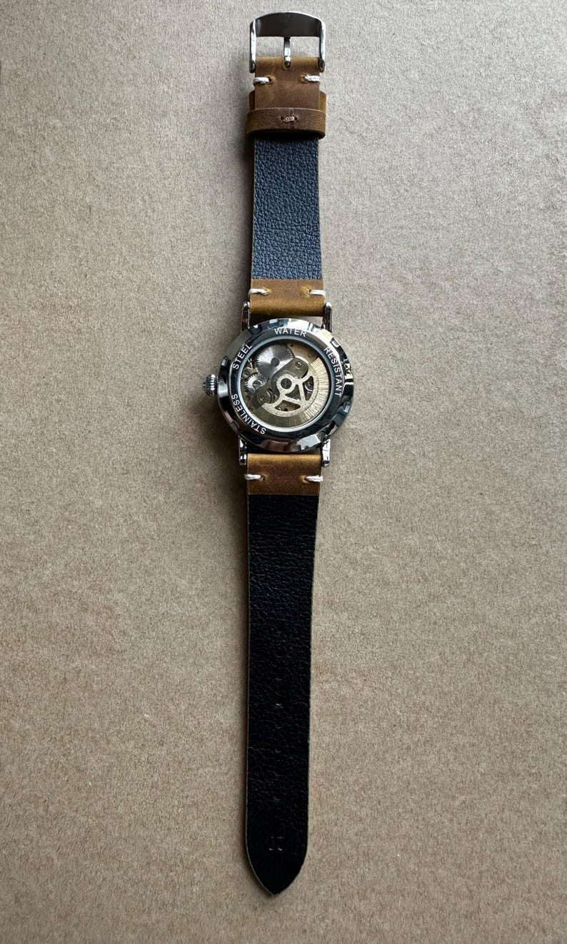 May include: A stainless steel wristwatch with a skeleton dial, showcasing the inner workings. The watch has a black leather strap with brown accents and a buckle closure. The watch face is visible, with the words "WATER RESISTANT" and "STAINLESS STEEL".