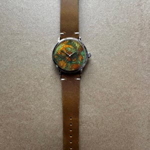 May include: A round wristwatch with a brown leather band. The watch face features a colorful fish design with shades of orange, green, and yellow. The watch has a silver-colored metal case and a buckle closure. The band is approximately 0.75 inches wide.