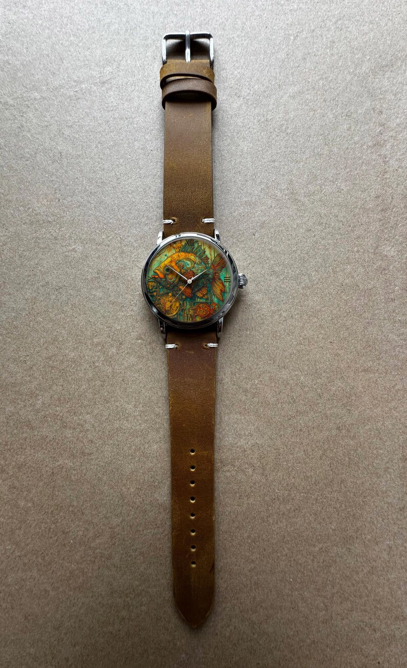 May include: A round wristwatch with a brown leather band. The watch face features a colorful fish design with shades of orange, green, and yellow. The watch has a silver-colored metal case and a buckle closure. The band is approximately 0.75 inches wide.