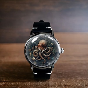 May include: A round wristwatch with a black leather band and a silver-colored case. The watch face features a detailed illustration of an octopus surrounded by plants. The watch has a domed crystal and white stitching on the band.