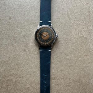 May include: A wristwatch with a navy blue leather band and a silver-colored case. The watch face features a detailed sunburst design in gold and brown tones. The band is approximately 1 inch wide.