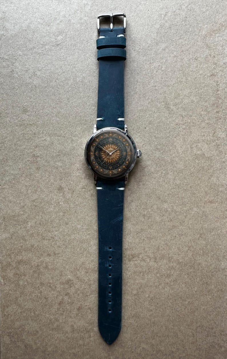 May include: A wristwatch with a navy blue leather band and a silver-colored case. The watch face features a detailed sunburst design in gold and brown tones. The band is approximately 1 inch wide.
