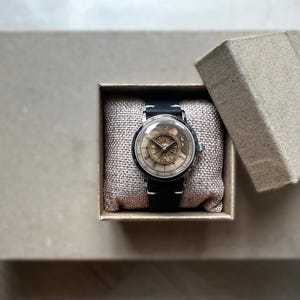 Puede incluir: Un reloj de estilo vintage con una correa de cuero negro y una caja plateada. La esfera del reloj presenta un dise&ntilde;o detallado de color dorado. Descansa dentro de una peque&ntilde;a caja de cart&oacute;n abierta sobre una superficie de tono neutro.