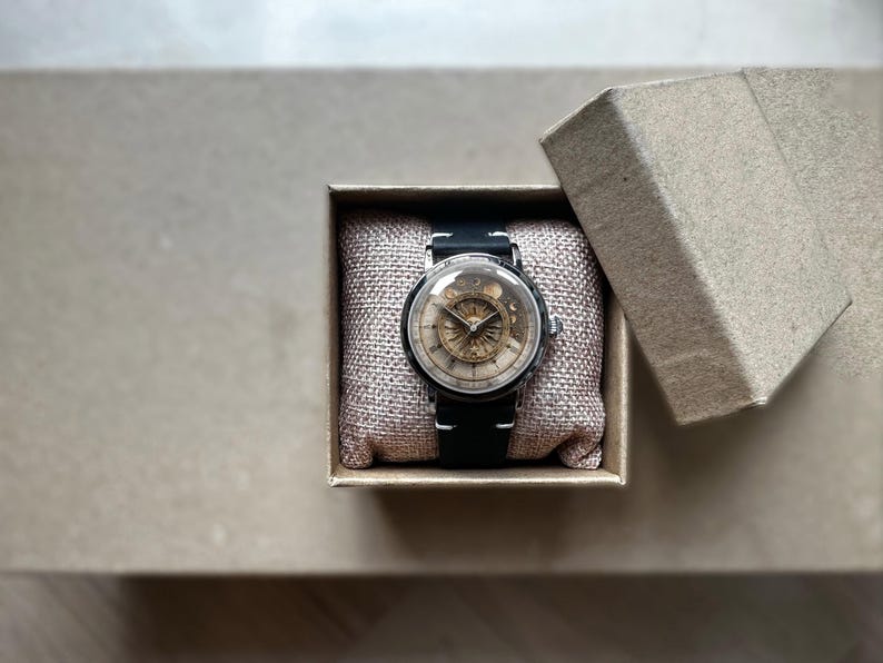 Puede incluir: Un reloj de estilo vintage con una correa de cuero negro y una caja plateada. La esfera del reloj presenta un dise&ntilde;o detallado de color dorado. Descansa dentro de una peque&ntilde;a caja de cart&oacute;n abierta sobre una superficie de tono neutro.