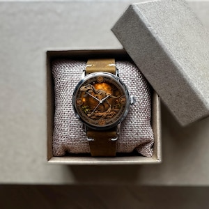 May include: A wristwatch with a brown leather band and a silver case. The watch face features a detailed illustration of a dinosaur. The watch is presented in a small, open, cardboard box, resting on a burlap pillow.