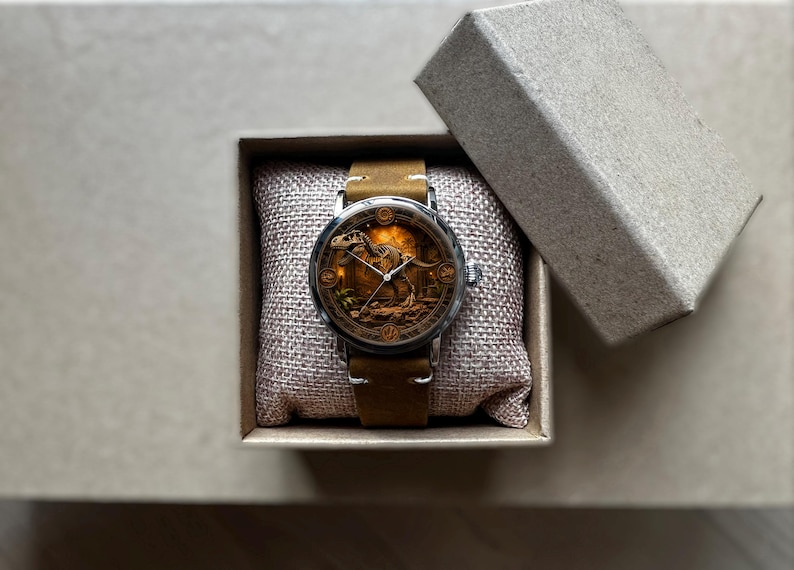 May include: A wristwatch with a brown leather band and a silver case. The watch face features a detailed illustration of a dinosaur. The watch is presented in a small, open, cardboard box, resting on a burlap pillow.