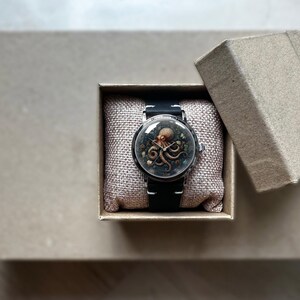 May include: A wristwatch with a black band and a round face featuring an octopus design. The watch is presented in an open, square, cardboard box with a linen-textured cushion. The octopus is brown and tan, set against a dark background.