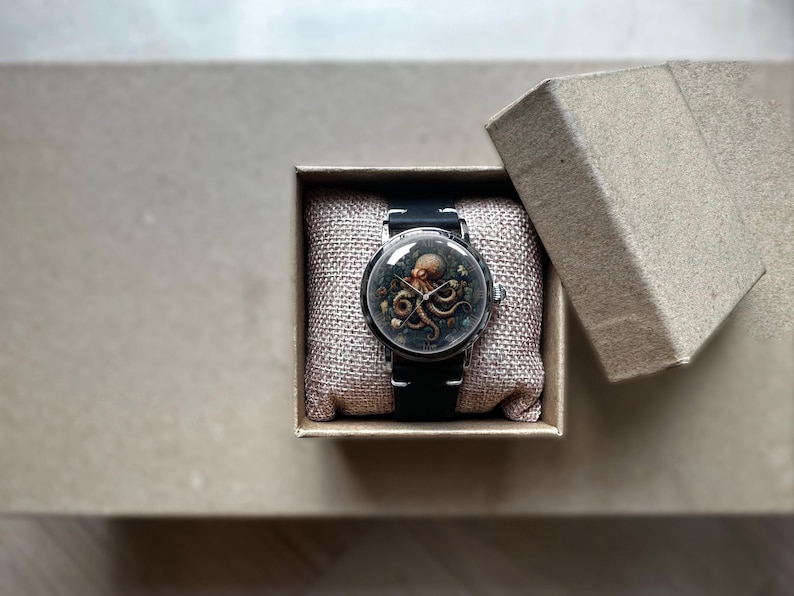 May include: A wristwatch with a black band and a round face featuring an octopus design. The watch is presented in an open, square, cardboard box with a linen-textured cushion. The octopus is brown and tan, set against a dark background.