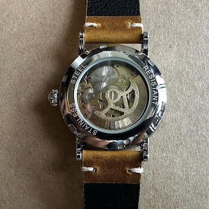May include: A stainless steel wristwatch with a transparent face revealing the intricate inner workings of the mechanism. The watch has a silver bezel, a tan leather strap, and a black leather backing. The watch is water resistant.