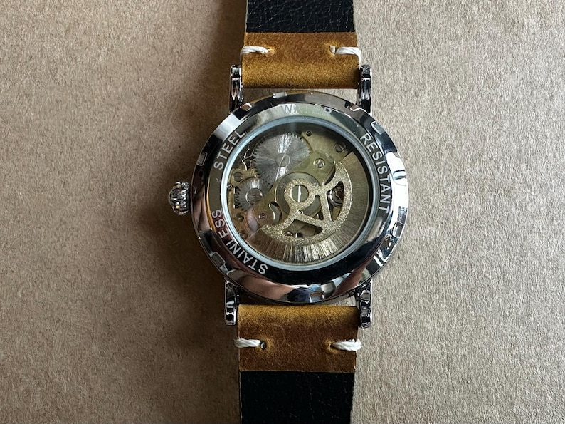 May include: A stainless steel wristwatch with a transparent face revealing the intricate inner workings of the mechanism. The watch has a silver bezel, a tan leather strap, and a black leather backing. The watch is water resistant.