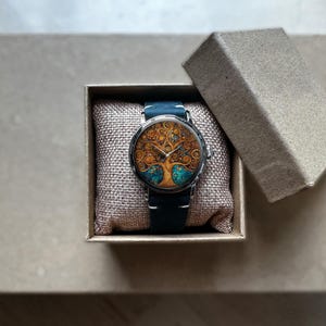 Puede incluir: Un reloj de pulsera redondo con un dise&ntilde;o detallado de &aacute;rbol de la vida en la esfera, con colores dorados y turquesa. El reloj tiene una correa de cuero azul oscuro y se presenta en una peque&ntilde;a caja de cart&oacute;n abierta. Un accesorio &uacute;nico.