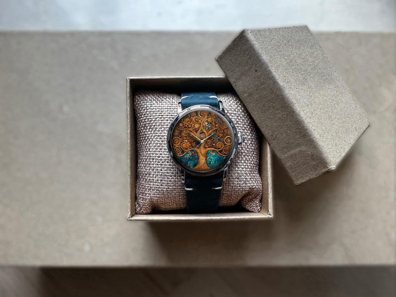 Puede incluir: Un reloj de pulsera redondo con un dise&ntilde;o detallado de &aacute;rbol de la vida en la esfera, con colores dorados y turquesa. El reloj tiene una correa de cuero azul oscuro y se presenta en una peque&ntilde;a caja de cart&oacute;n abierta. Un accesorio &uacute;nico.