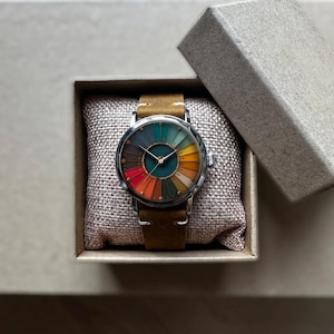 May include: A wristwatch with a colorful, segmented face, presented in a gift box. The watch features a brown leather strap and a silver-colored case. The face displays a spectrum of colors, including red, orange, yellow, green, and brown.