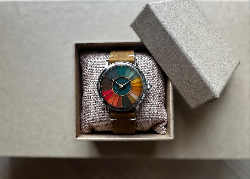 May include: A wristwatch with a colorful, segmented face, presented in a gift box. The watch features a brown leather strap and a silver-colored case. The face displays a spectrum of colors, including red, orange, yellow, green, and brown.