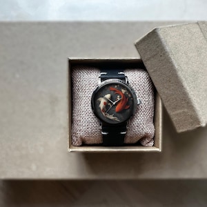 Op de afbeelding: Een ronde polshorloge met een zwarte band en een zilverkleurige kast, gepresenteerd in een kleine, open kartonnen doos. De wijzerplaat heeft een koi-vissenontwerp in tinten rood, wit en zwart. Het horloge ligt op een beige kussen.