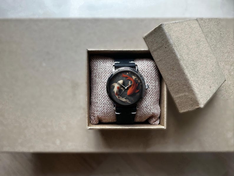 Op de afbeelding: Een ronde polshorloge met een zwarte band en een zilverkleurige kast, gepresenteerd in een kleine, open kartonnen doos. De wijzerplaat heeft een koi-vissenontwerp in tinten rood, wit en zwart. Het horloge ligt op een beige kussen.