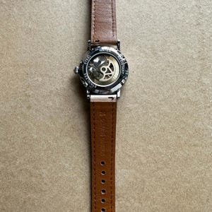 May include: A stainless steel watch with a brown leather strap. The watch face is transparent, revealing the inner workings. The strap is secured with a silver buckle and has multiple adjustment holes. The watch is water resistant.