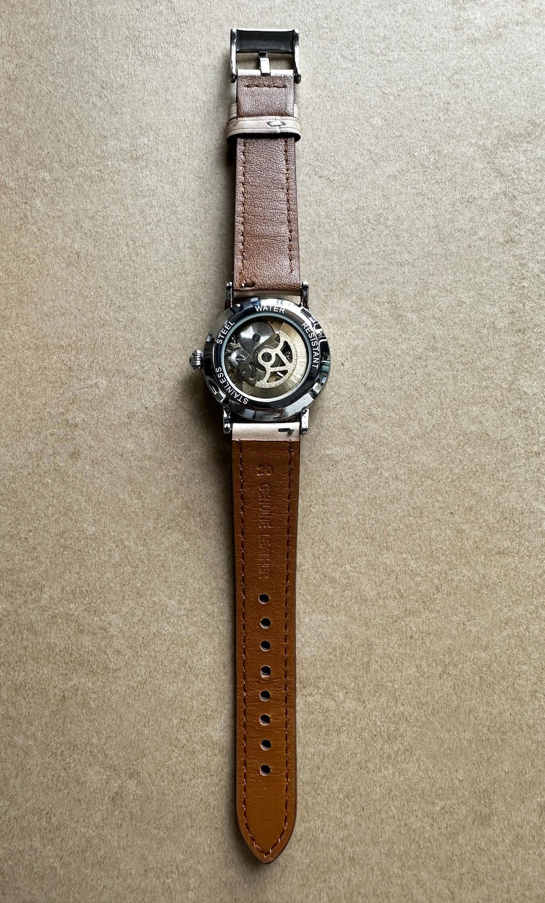 May include: A stainless steel watch with a brown leather strap. The watch face is transparent, revealing the inner workings. The strap is secured with a silver buckle and has multiple adjustment holes. The watch is water resistant.