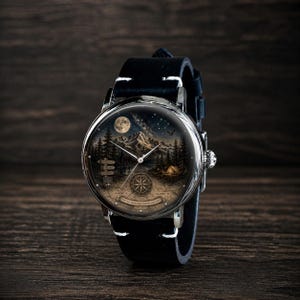 May include: A round wristwatch with a black leather band. The watch face features a detailed landscape with mountains, trees, a full moon, and a compass design. The watch has silver-colored metal casing.