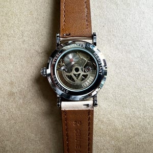 May include: A wristwatch with a brown leather band and a silver-colored stainless steel case. The watch face is visible, showcasing the intricate mechanical movement. The back of the watch is marked "Water Resistant" and "Stainless Steel".