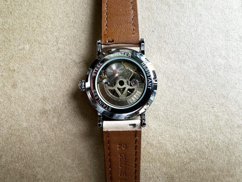 May include: A wristwatch with a brown leather band and a silver-colored stainless steel case. The watch face is visible, showcasing the intricate mechanical movement. The back of the watch is marked "Water Resistant" and "Stainless Steel".